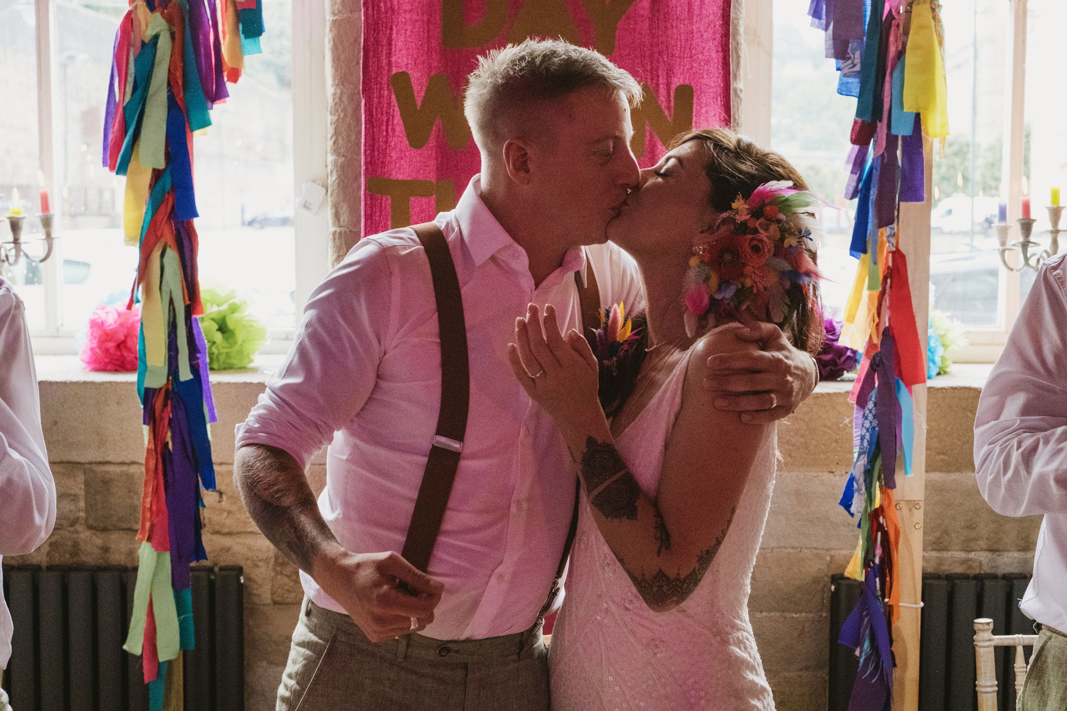 Couple kissing beneath colourful ribbons during lively wedding celebration, arms wrapped close.