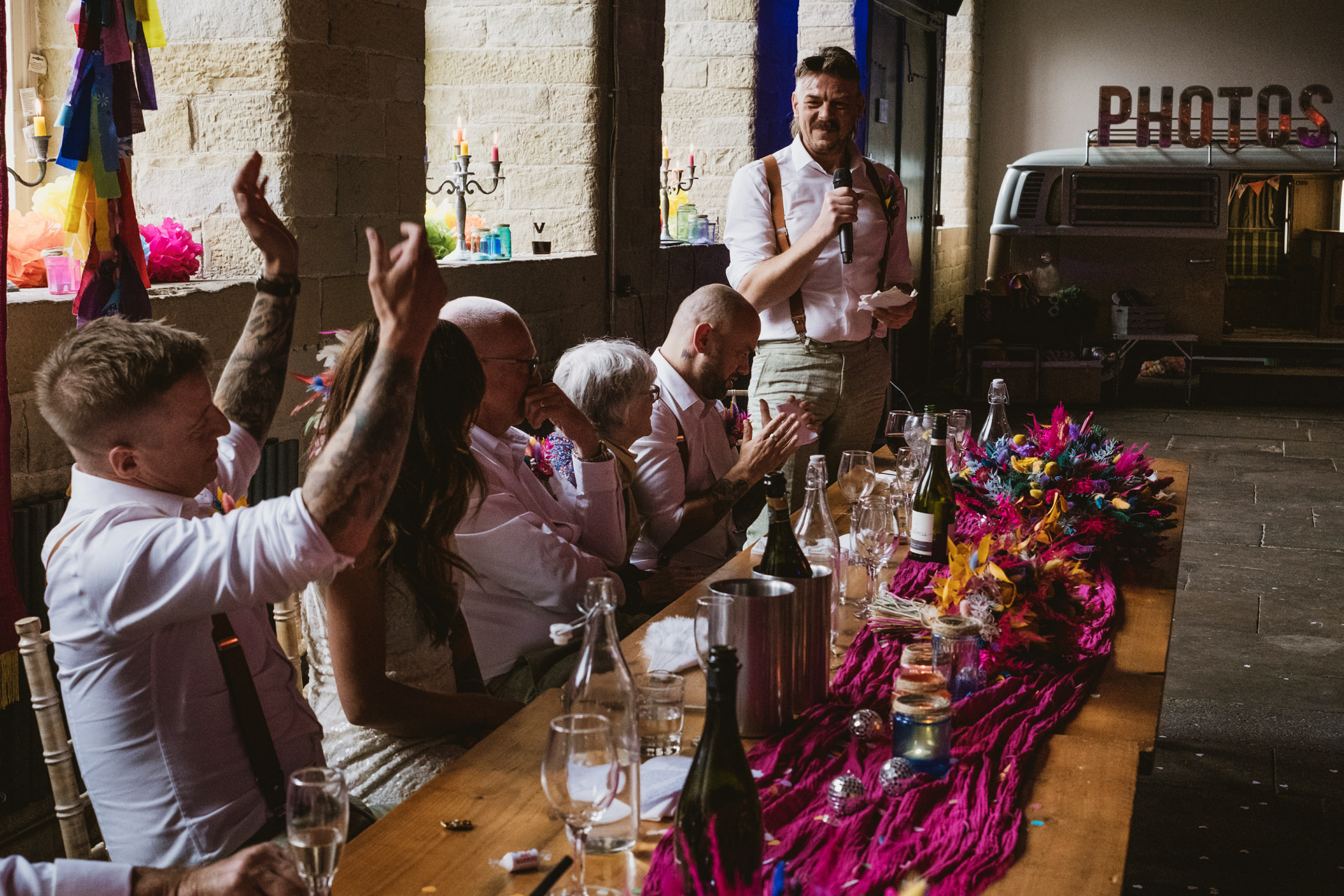 Wedding speech during lively reception, guests raising hands and laughing around candlelit banquet table.
