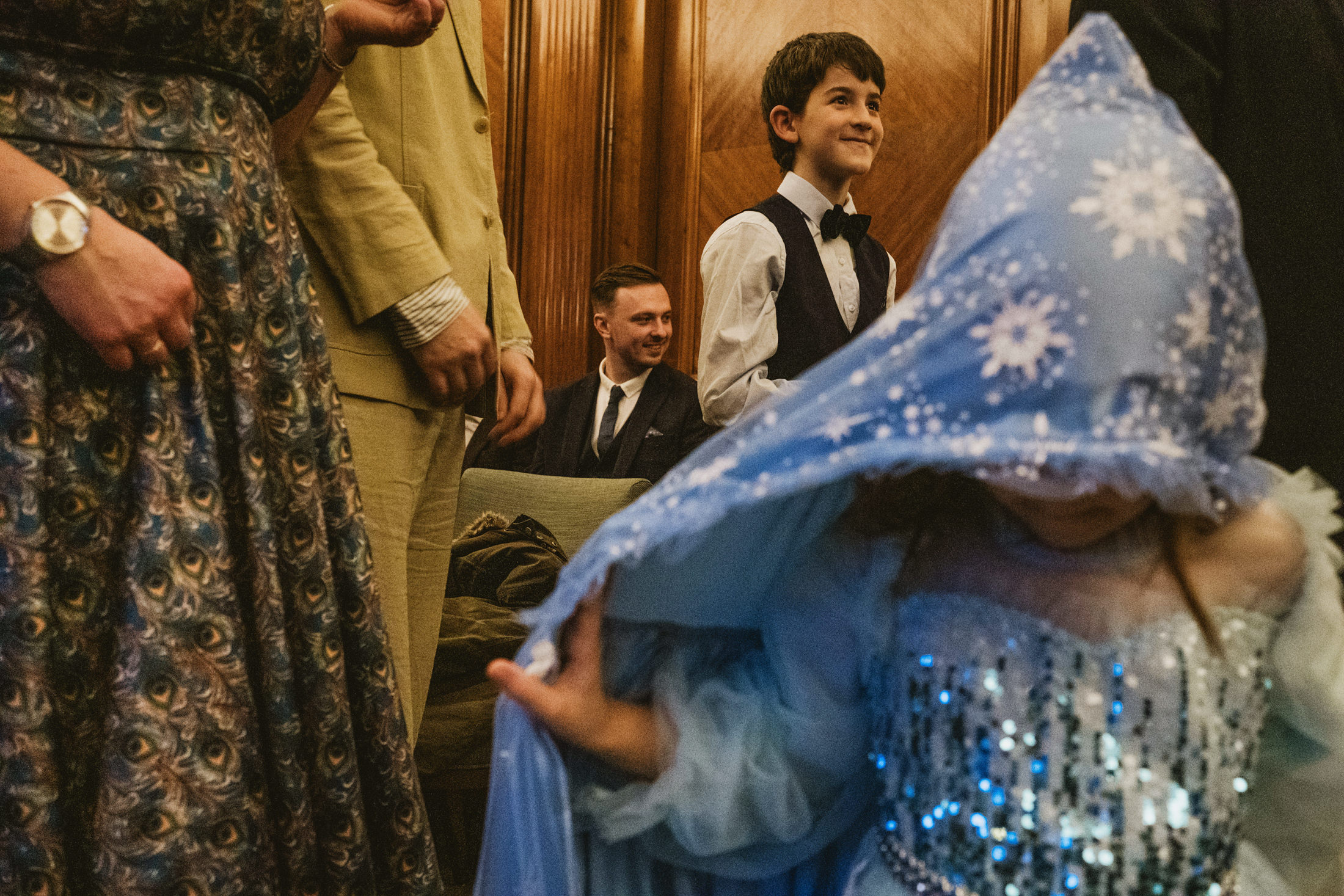 Child in blue sequinned cape twirls as boy smiles during wedding ceremony, guests watching warmly.
