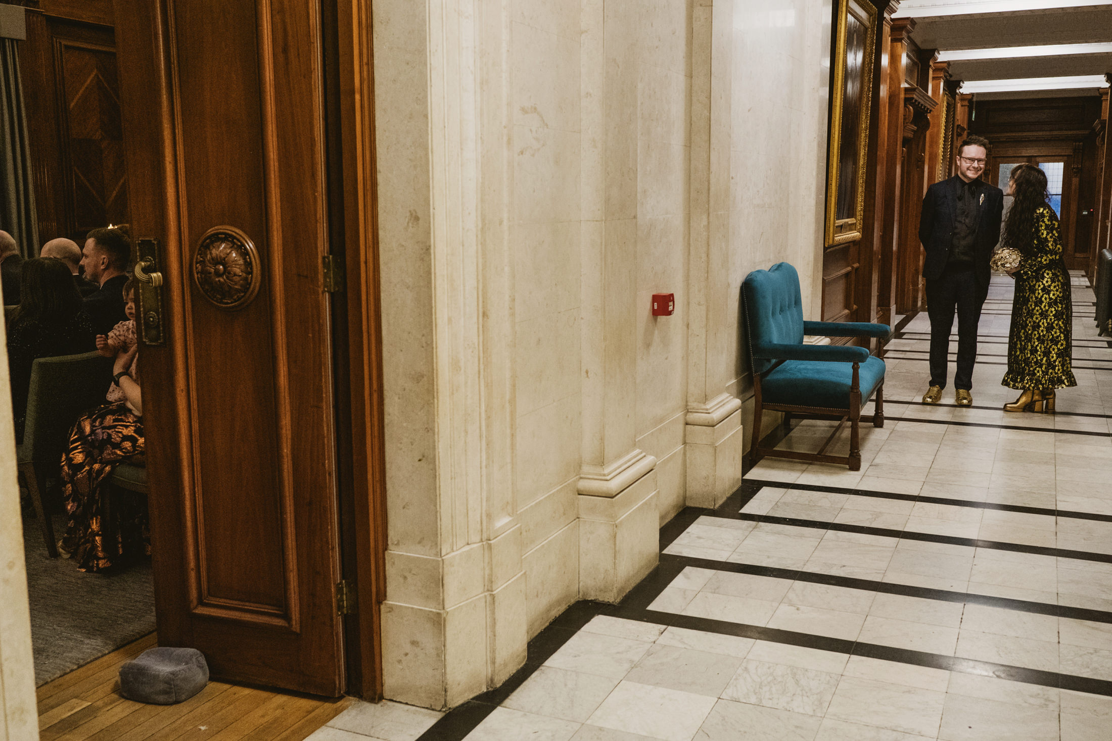 Couple smiling in marble hallway as guests gather beyond wooden doors during wedding photography.