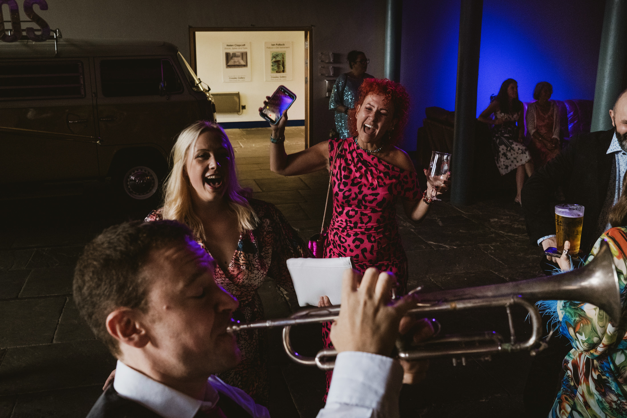 Wedding guests laugh and dance as a trumpeter plays, phone raised, wine glass glowing in blue light.