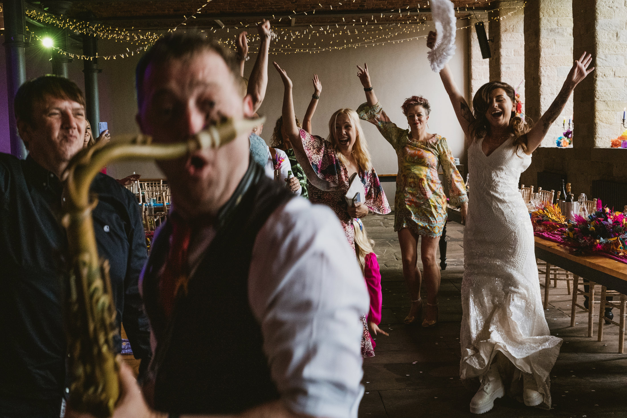Bride and guests cheer as saxophonist plays during lively wedding reception, arms raised beneath twinkling lights.