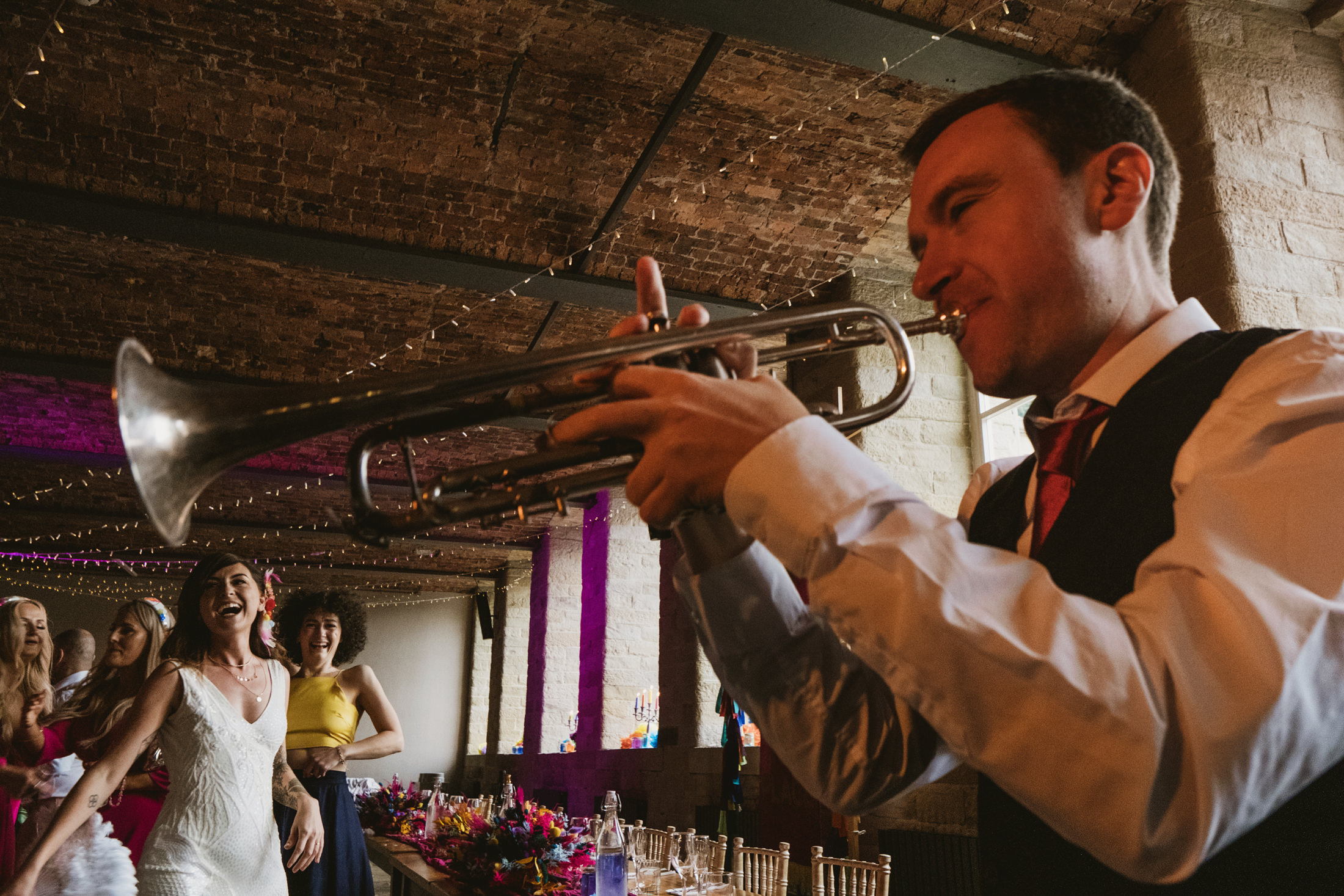 Trumpeter playing as guests laugh and dance beneath fairy lights at rustic wedding reception